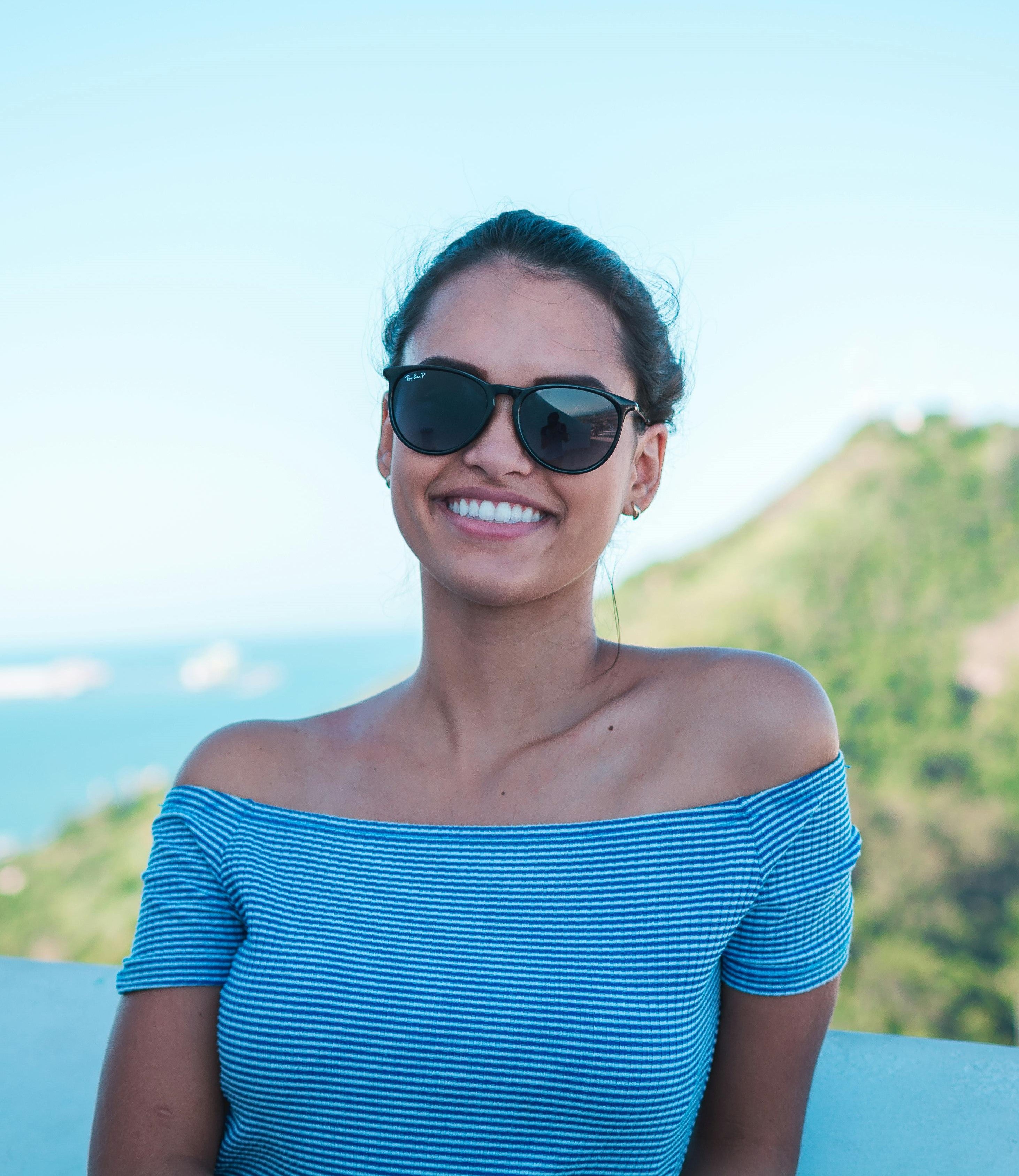 Brown Woman on holiday wearing black prescription sunglasses taking a picture in front of big green mountains and the sea.